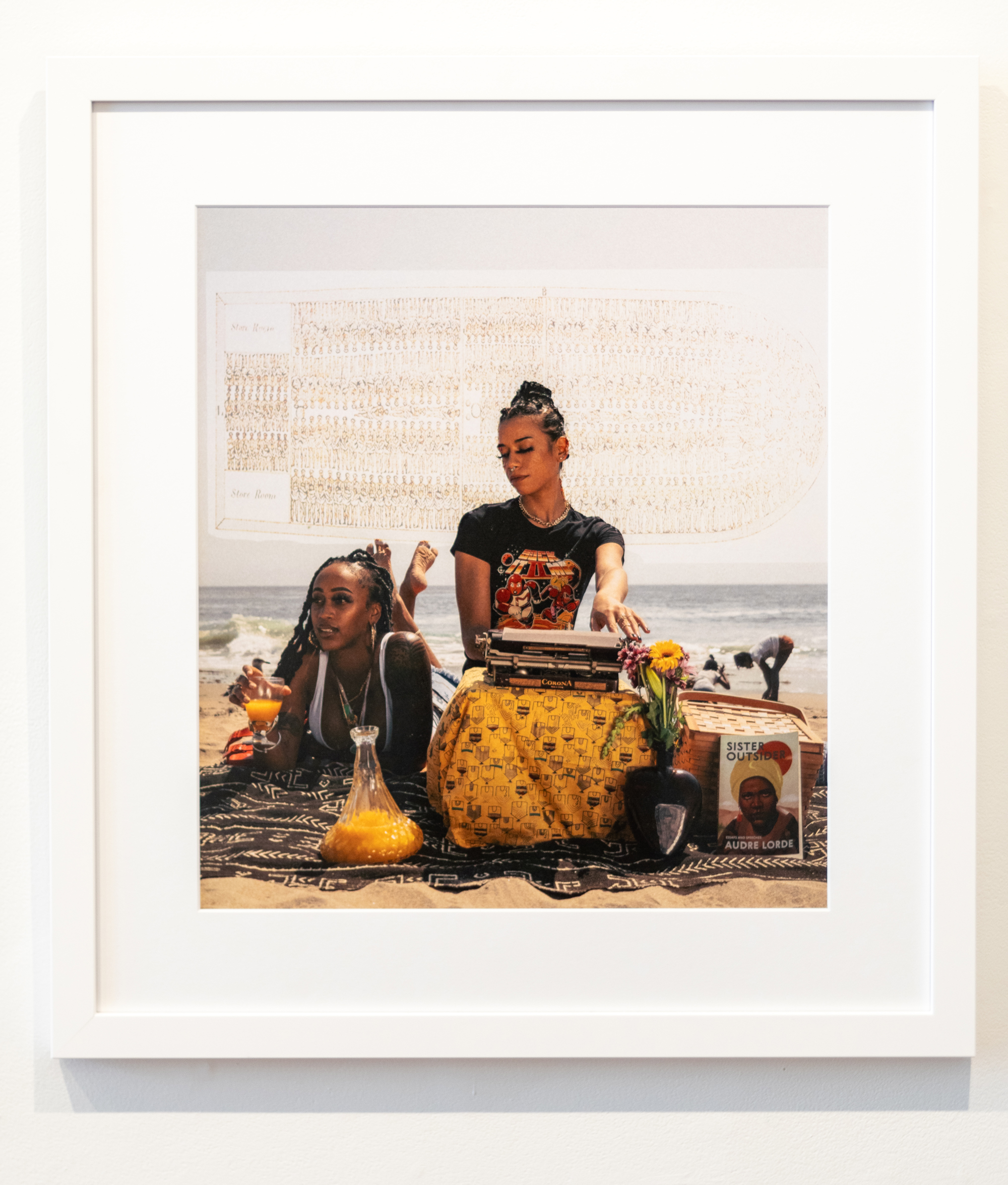 Framed color photo of two women at beach with drawing of slave ship behind them