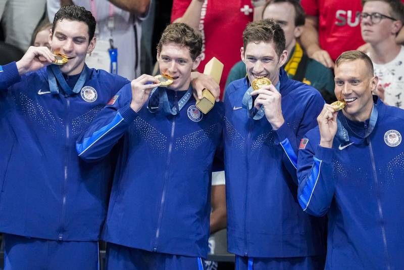 Four white men in blue tracksuits stand in a line each biting their own gold medals.