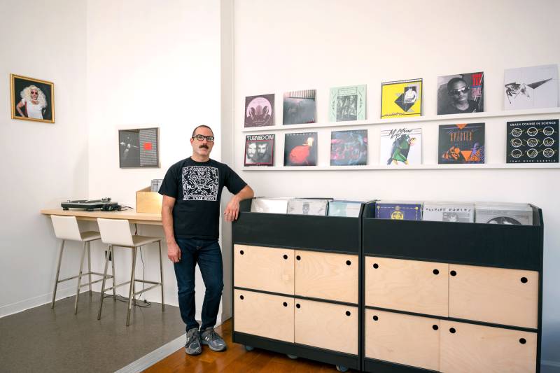 Man stands leaning on bins of records with display of covers on wall behind