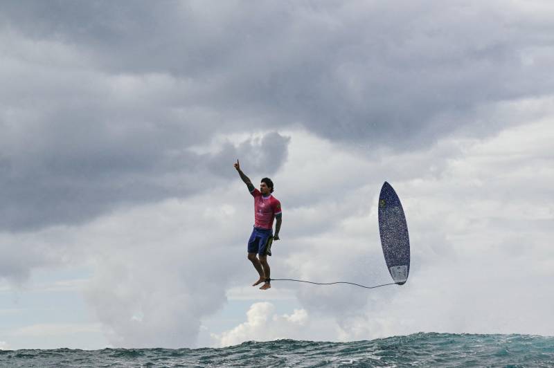 A surfer stands upright in midair, with one arm extended aloft. Behind him trails his surfboard, tethered to his ankle, also in midair. The ocean waves roll several feet below.