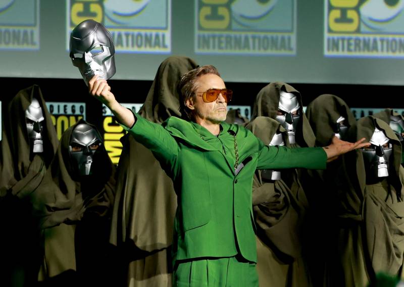 A man wearing a striking green suit stands arms extended, holding up a silver mask.