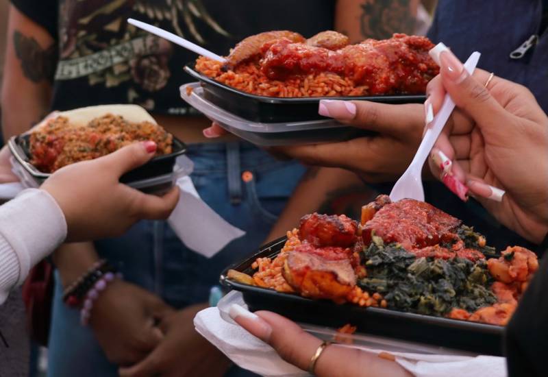 Customers eating jollof rice out of black plastic takeout containers.