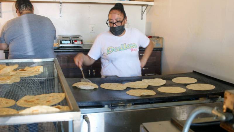 A woman in a black face mask flips flour tortillas cooking on a griddle.