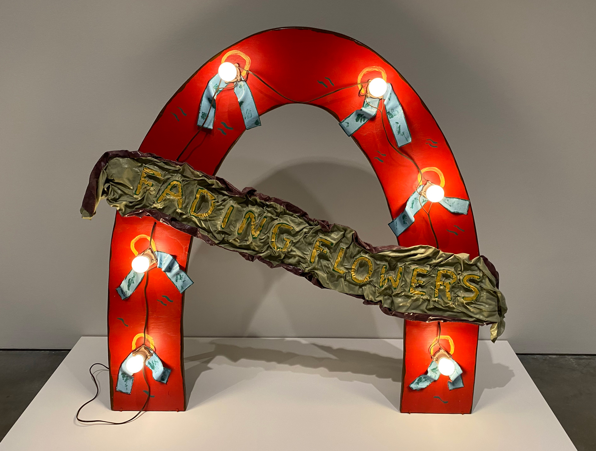 Red horseshoe-shaped sculpture with light bulbs and a banner reading FADING FLOWERS