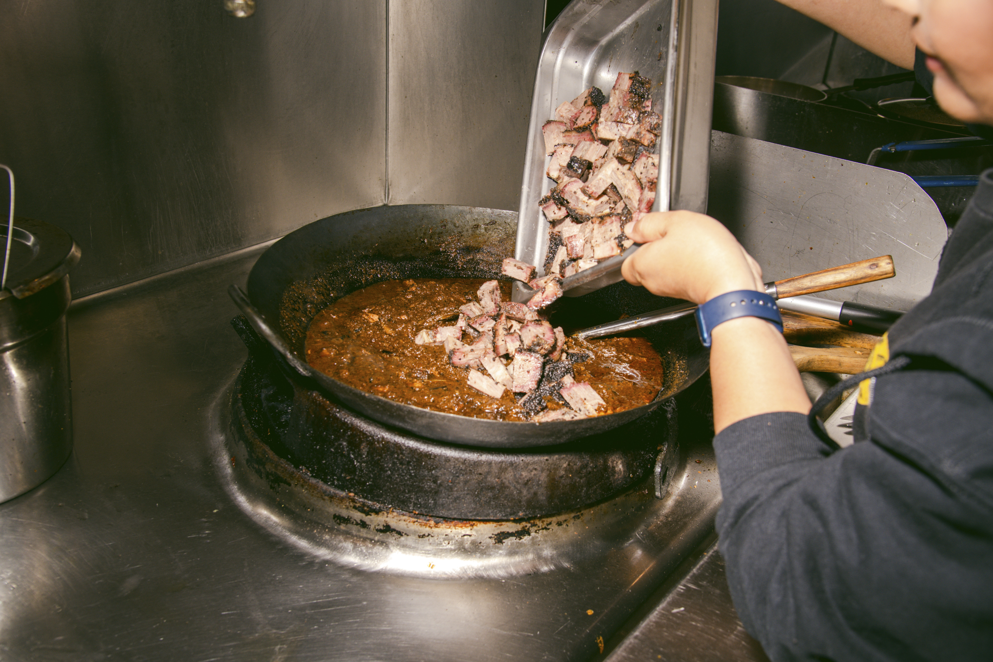 A chef dumps a tray of cubed brisket into a pot of brown stew.