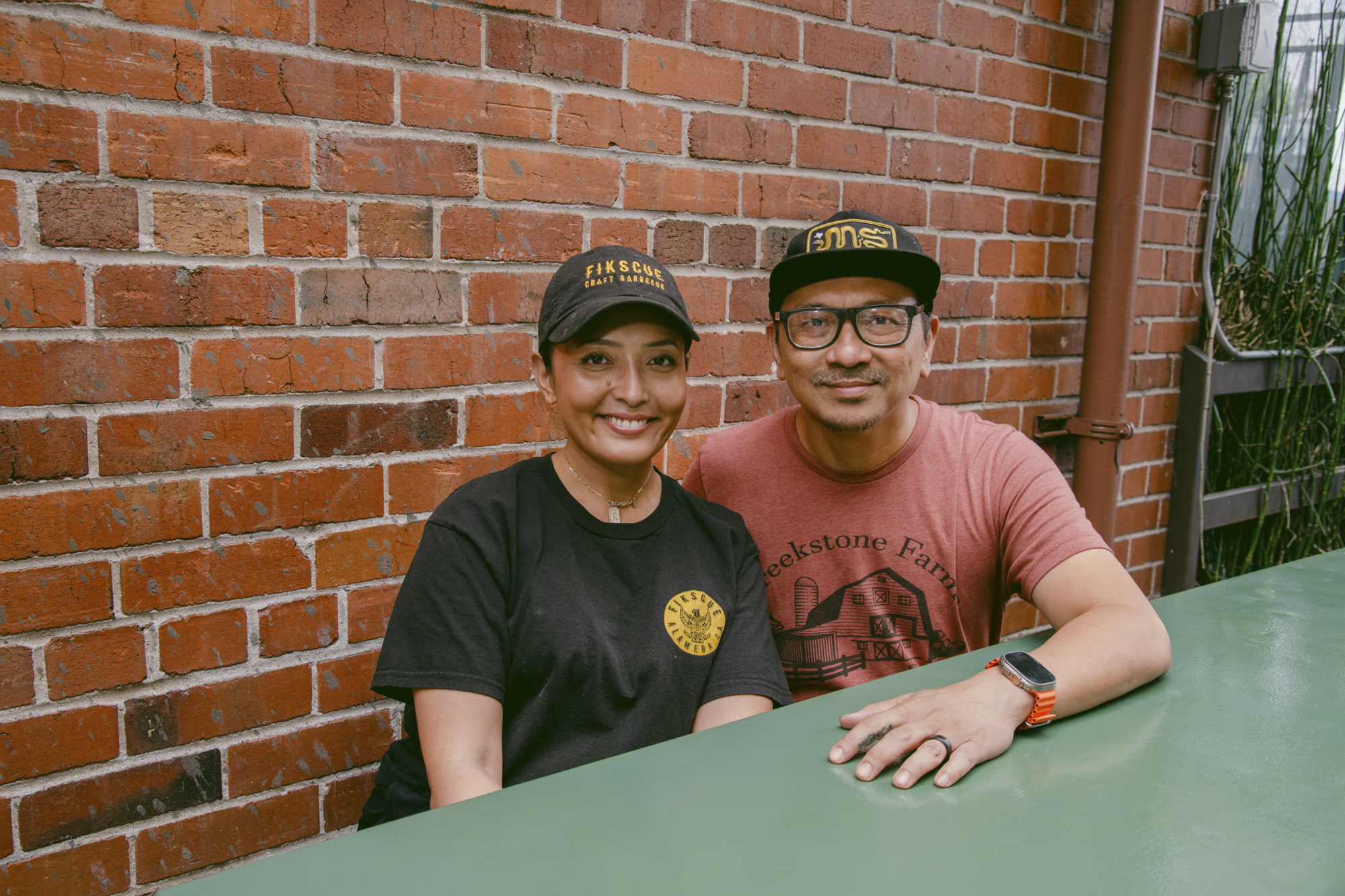 A woman and a man pose for a portrait, both wearing black and gold "Fikscue" baseball caps.