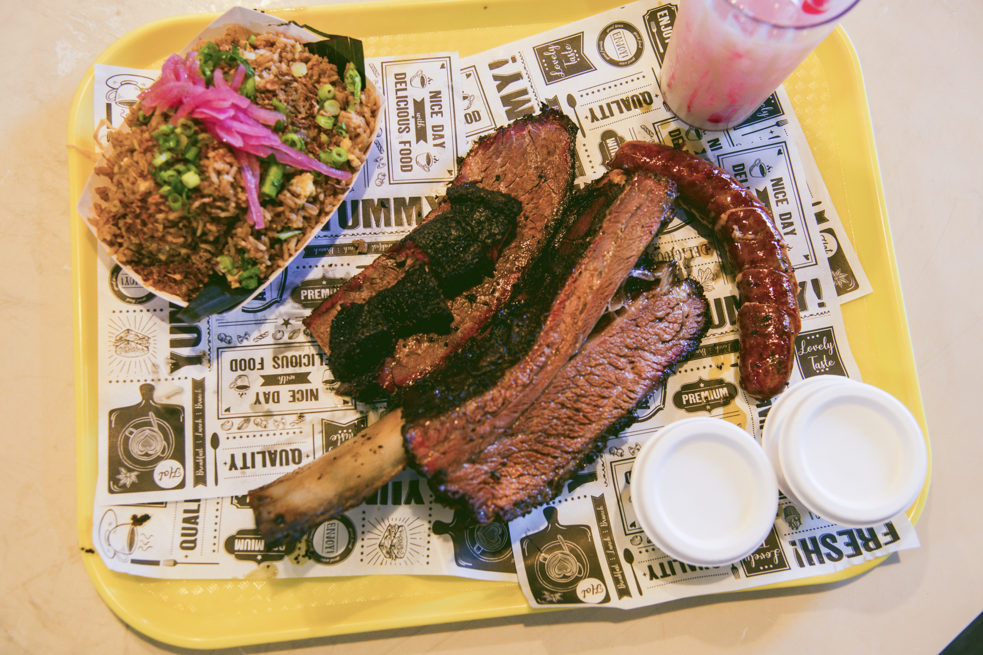 A tray of barbecue includes a dino beef rib, a slice of brisket, a sausage, and a cardboard try of fried rice.