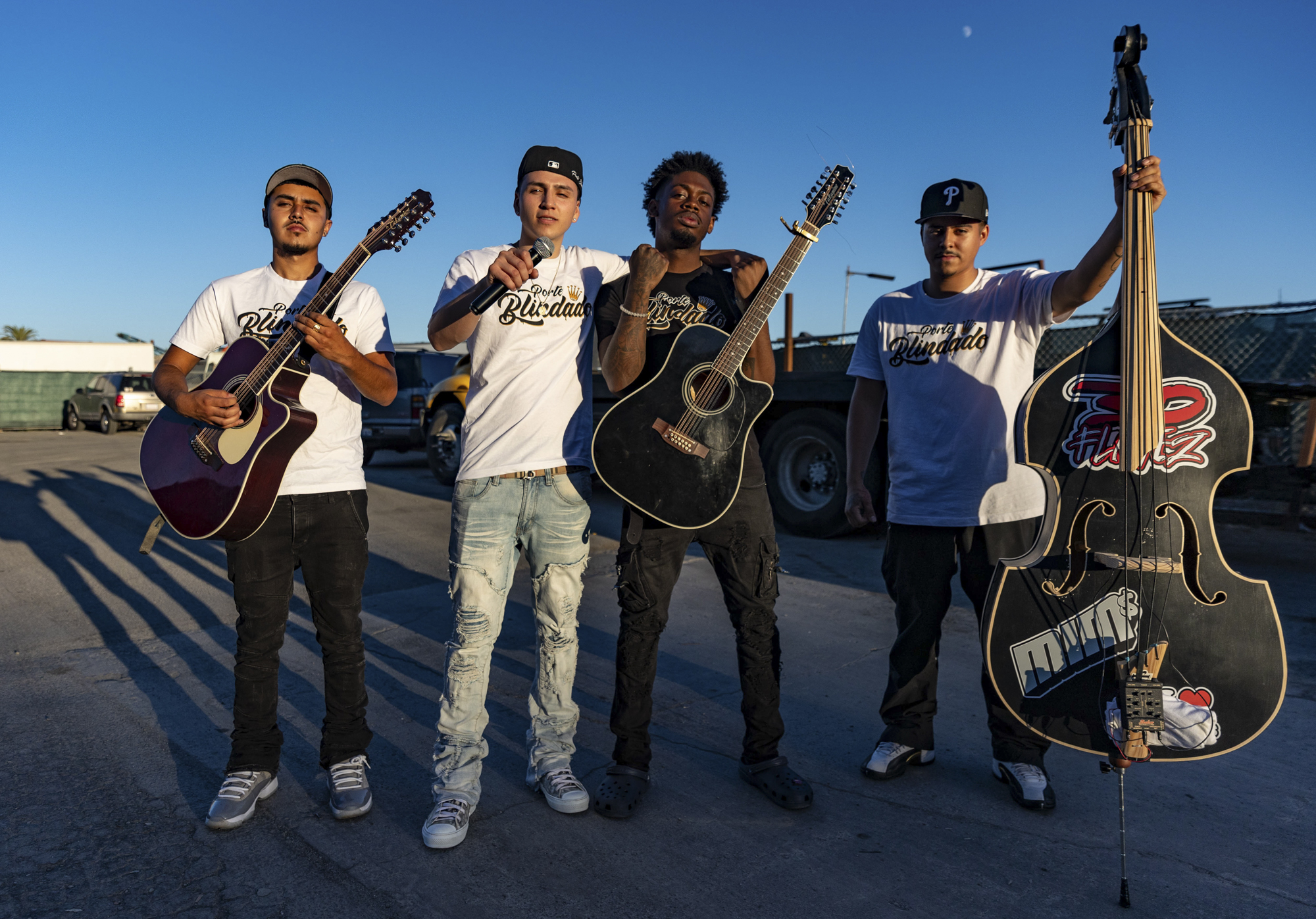 Four young musicians pose with their instruments in an industrial parking lot at sunset.