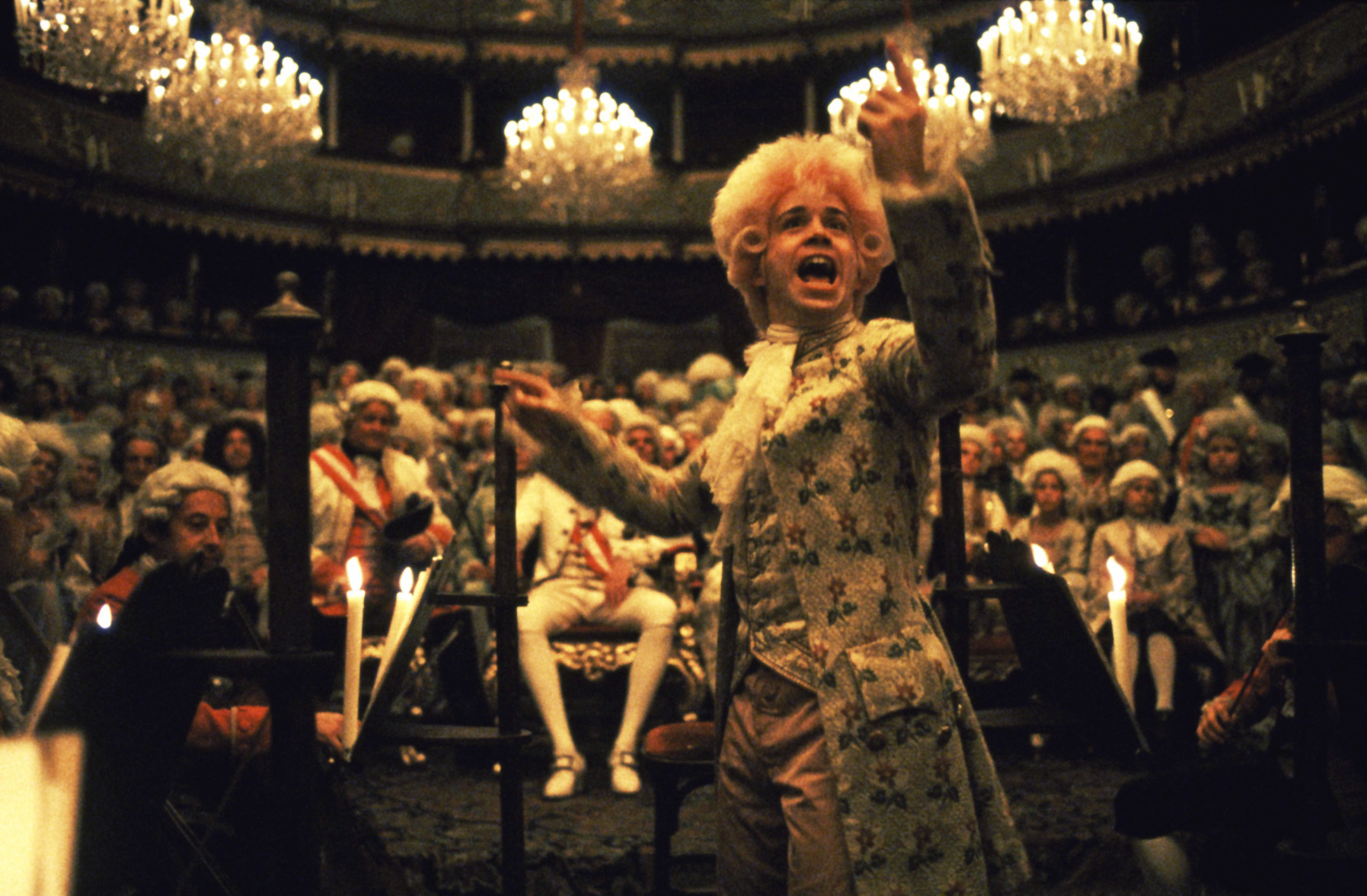 Man in baroque clothing and wig gesture broadly in concert hall