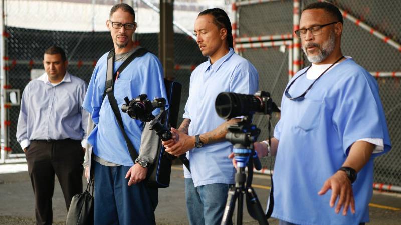 Three men in blue prison wear line up a shot with cameras.