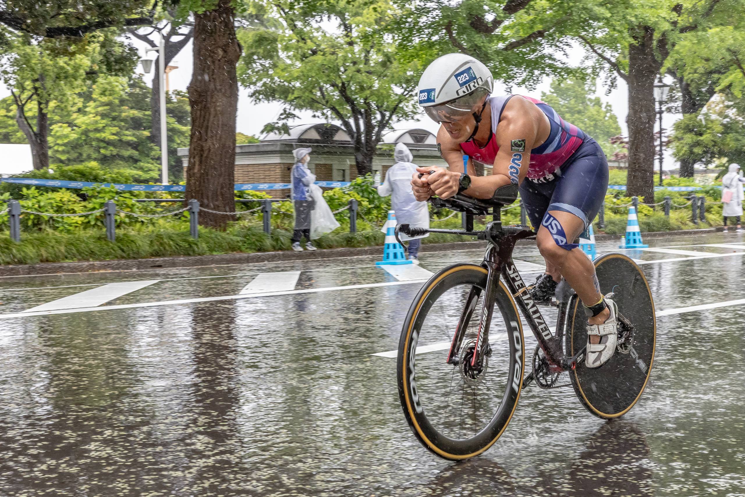 A competitive cyclist in full racing gear rides down a rain-drenched road.