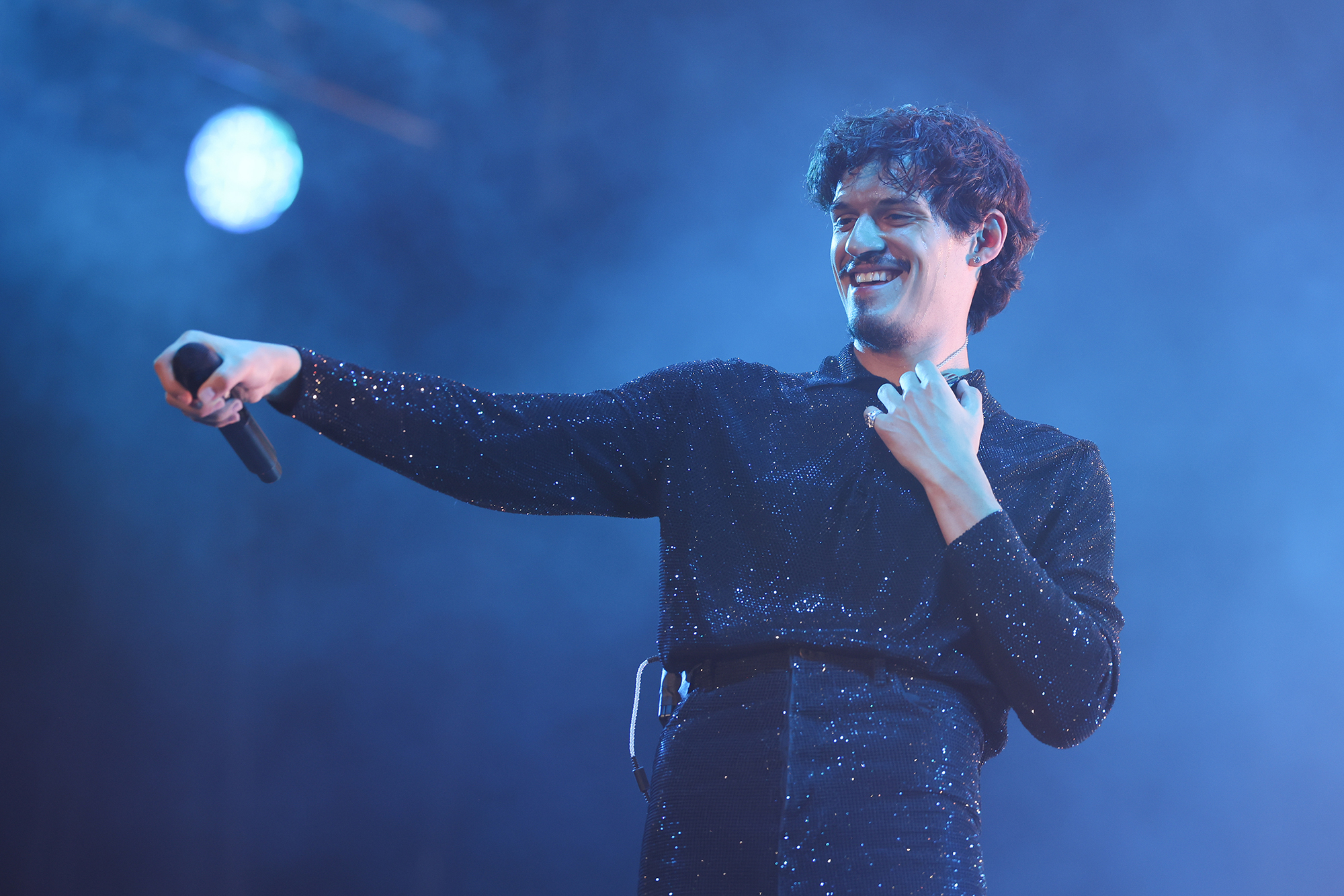 Omar Apollo smiles and looks out onto the crowd while holding a microphone and wearing sparkly black pants and a matching top. 