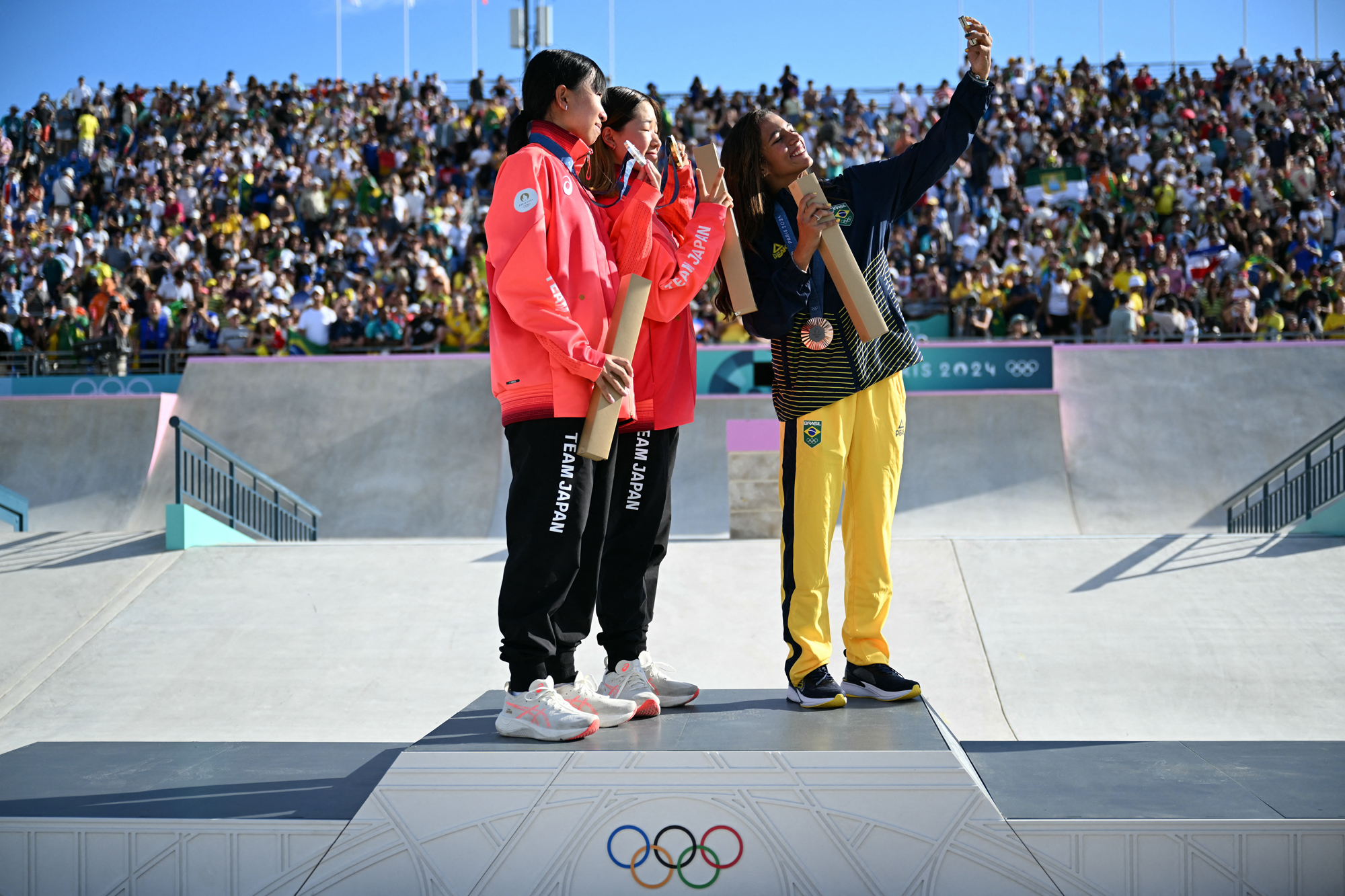 Three young women gather together and smile for one of their cell phones, held aloft, while atop an Olympic podium, watched by large crowds in the distance.