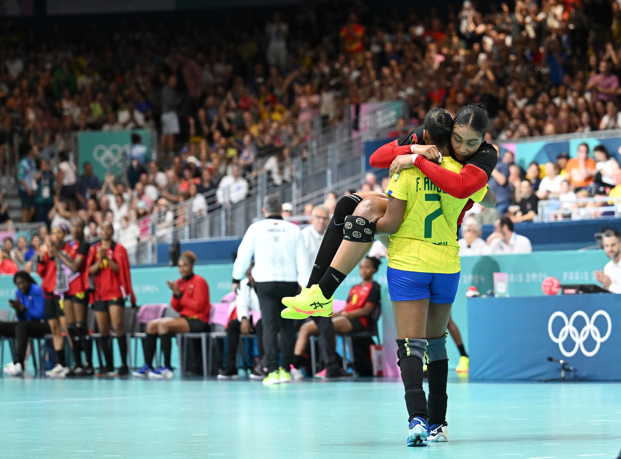 Large crowds watch on as a female handball player carries a rival handball player off the court after an injury.