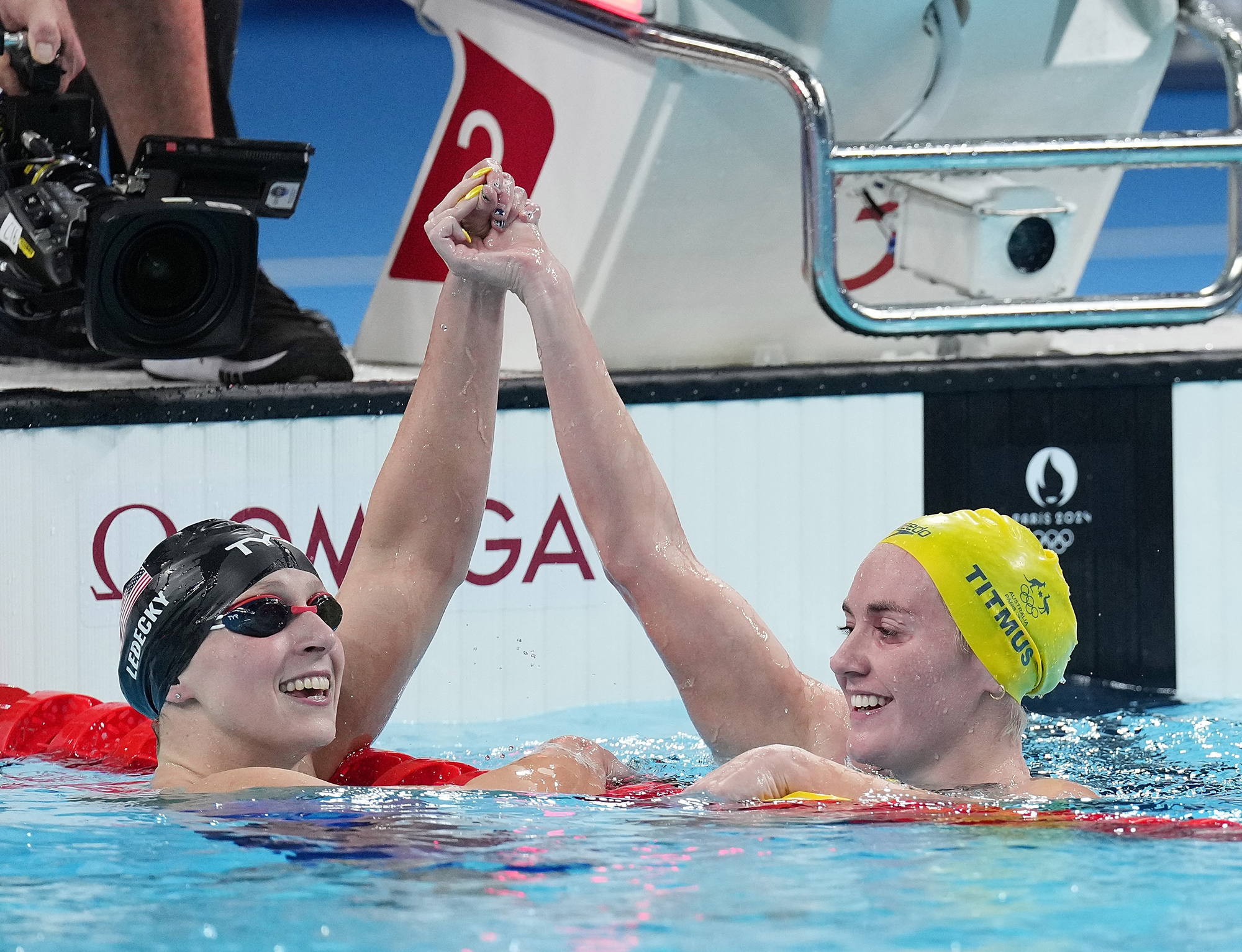 Smiling female swimmers hold hands with their arms raised high across a pool lane divider.