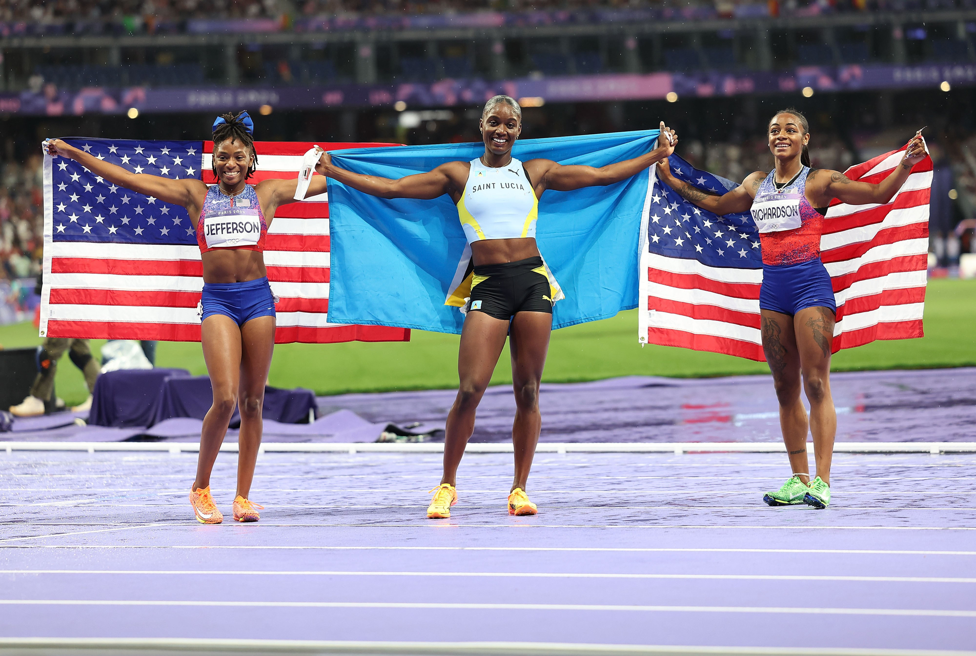 Three female athletes smile triumphantly on a running track holding up their countries' flags.