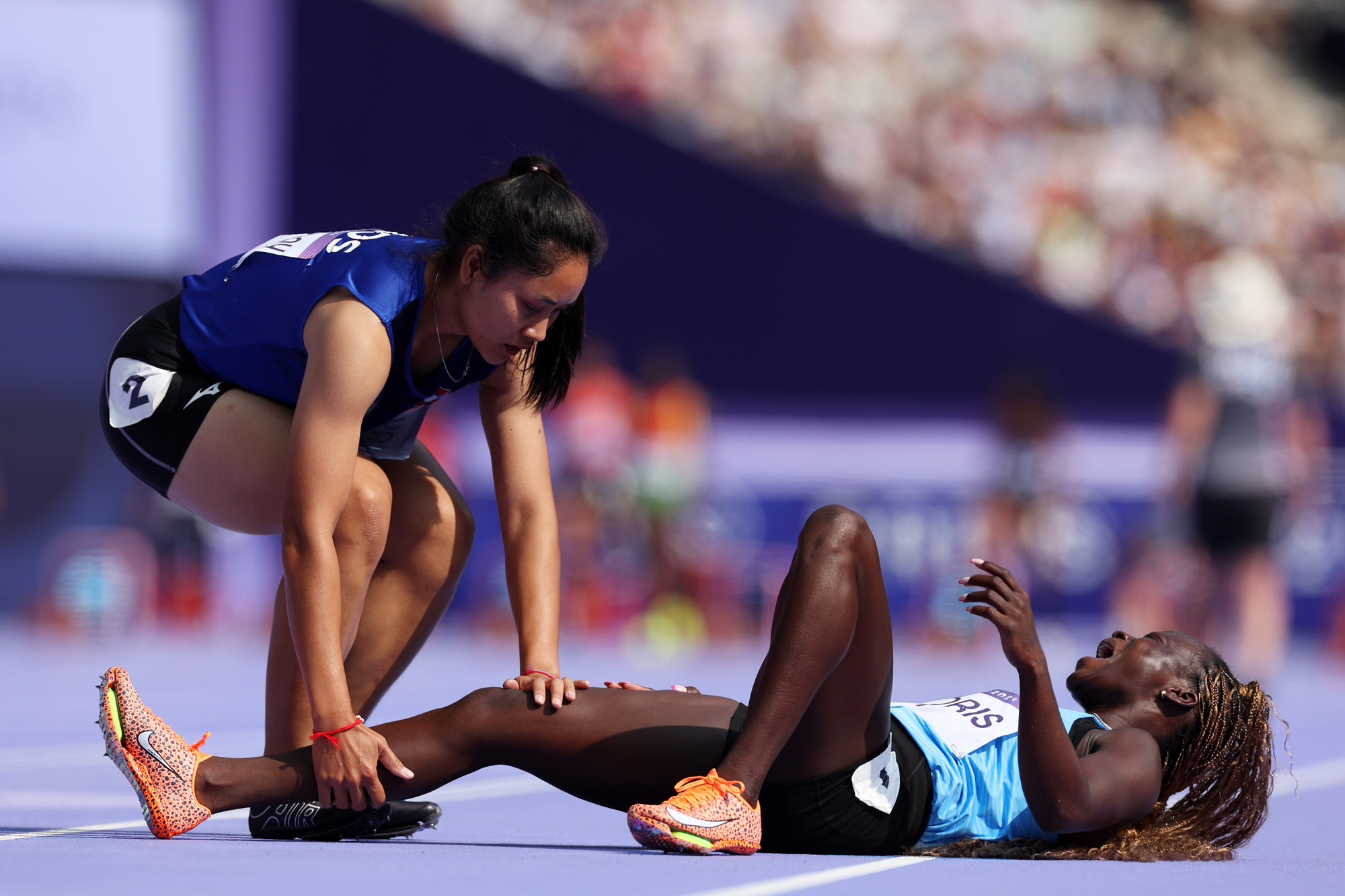 One female runner lies on the track distressed, while another bends down and tries to support the injured runner's leg.