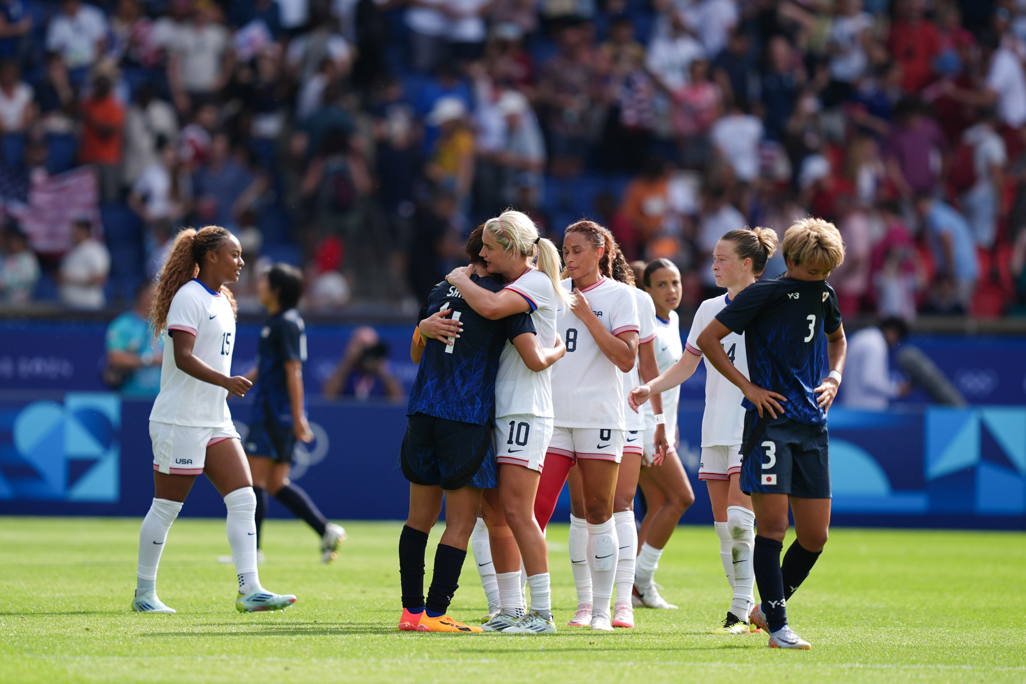 A woman in a white soccer uniform hugs a woman in black and blue soccer uniform on a field in front of a packed crowd.