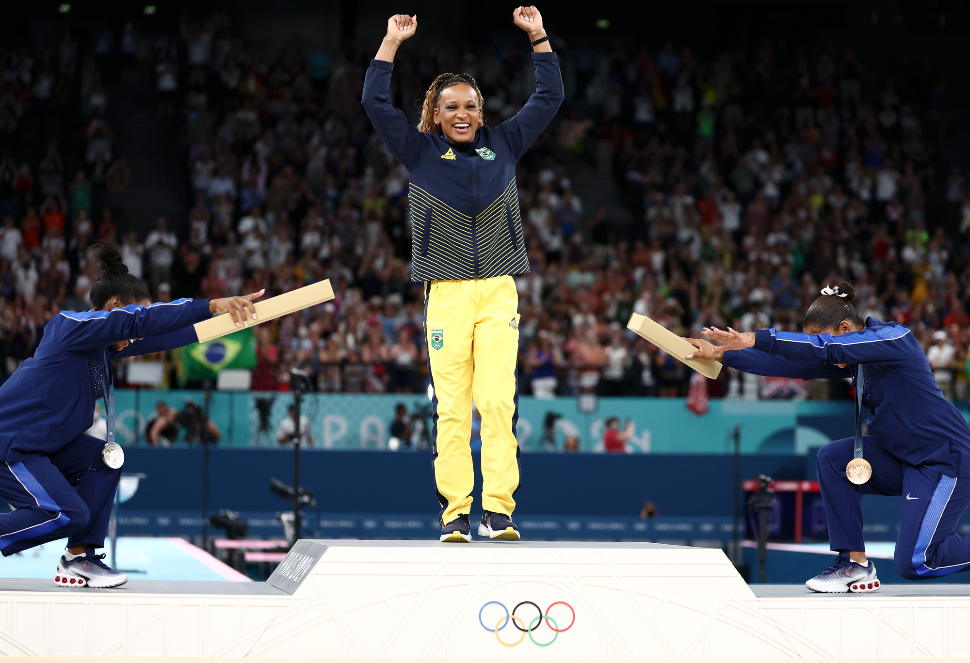 An athlete stands arms raised and triumphant on a podium, two fellow athletes bow down in praise on either side