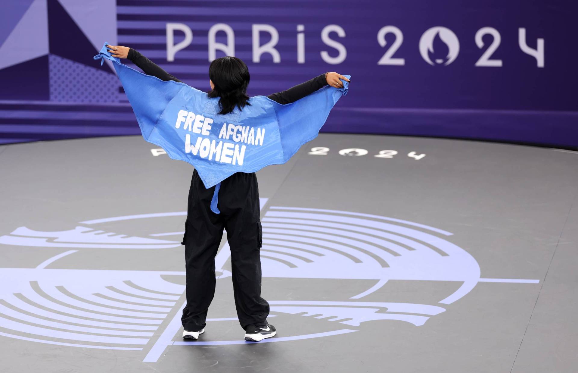 An athlete on an Olympic stage stands with her arms outstretched wearing a blue cape that says "Free Afghan Women."