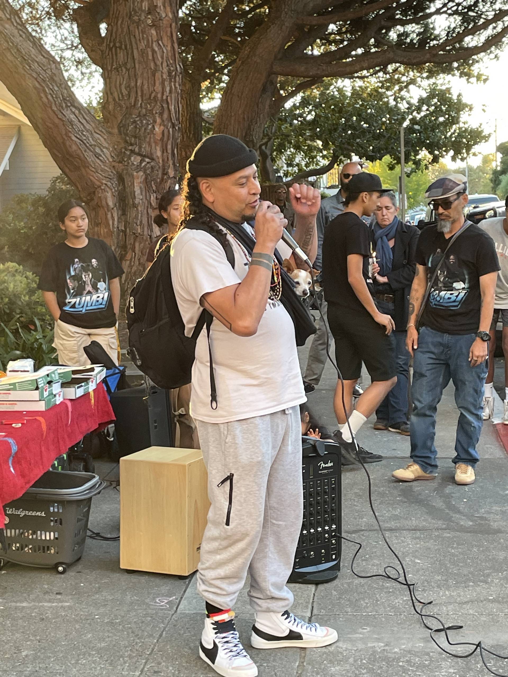 A man speaks into the microphone at a small gathering on the sidewalk. 