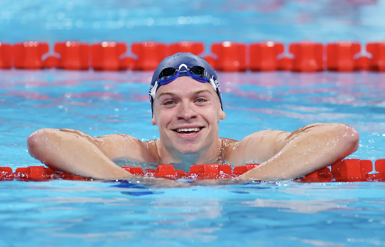 A male Olympic swimmer props himself up on a lane divider in a swimming pool and smiles broadly.