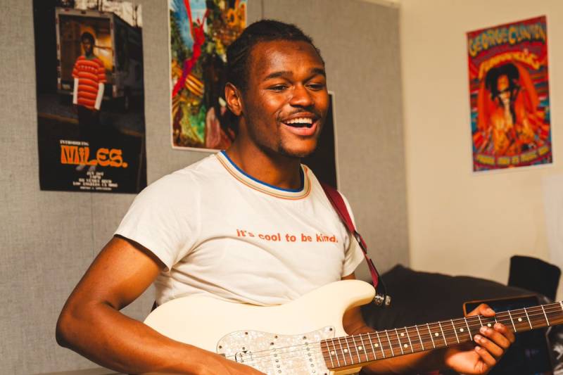 A young Black man sits on a bed with posters on the wall in the background, playing a guitar in a white t-shirt that reads 'it's cool to be kind'
