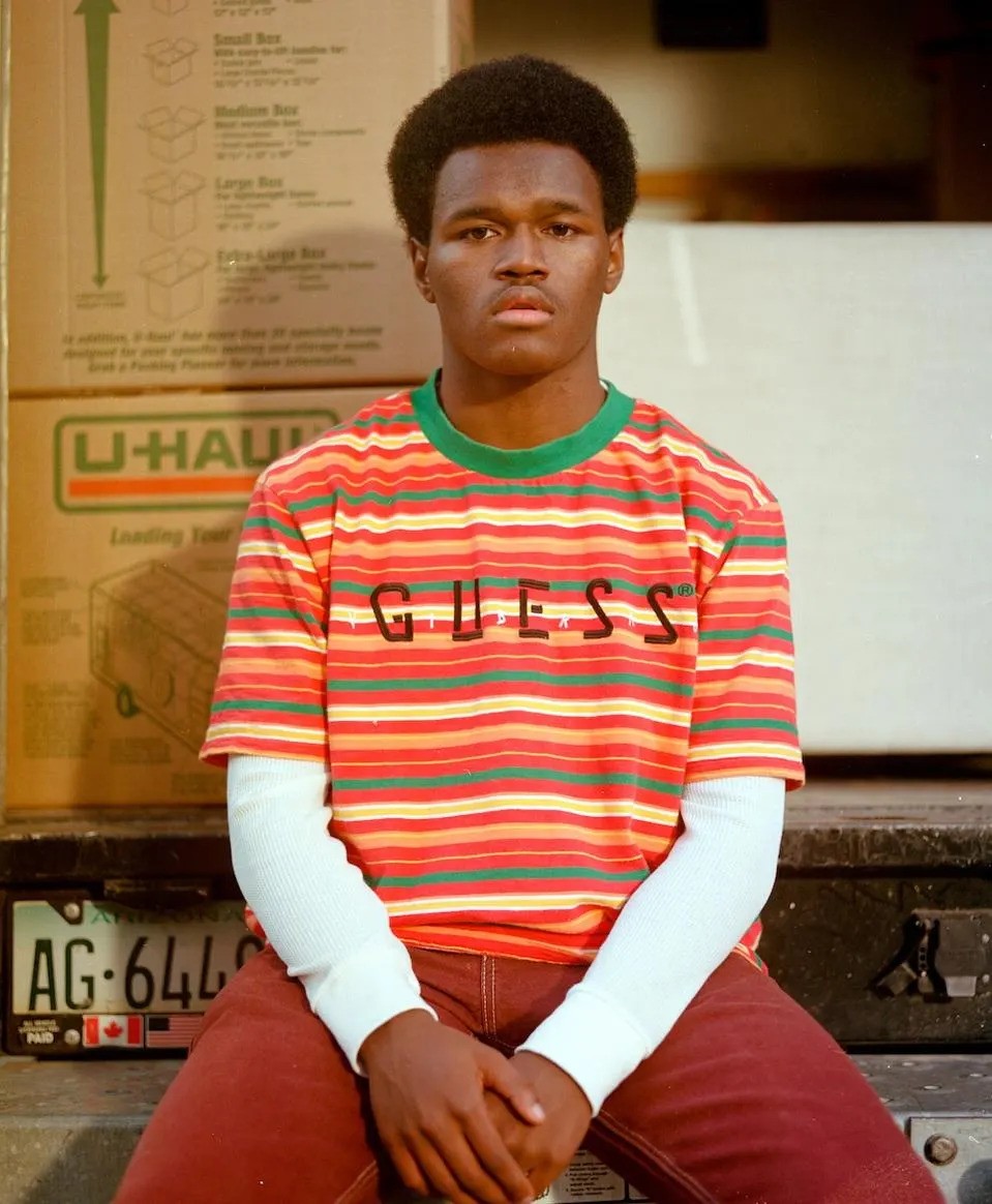A young Black man in a multicolored striped Guess brand shirt sits on the back of a U-Haul truck