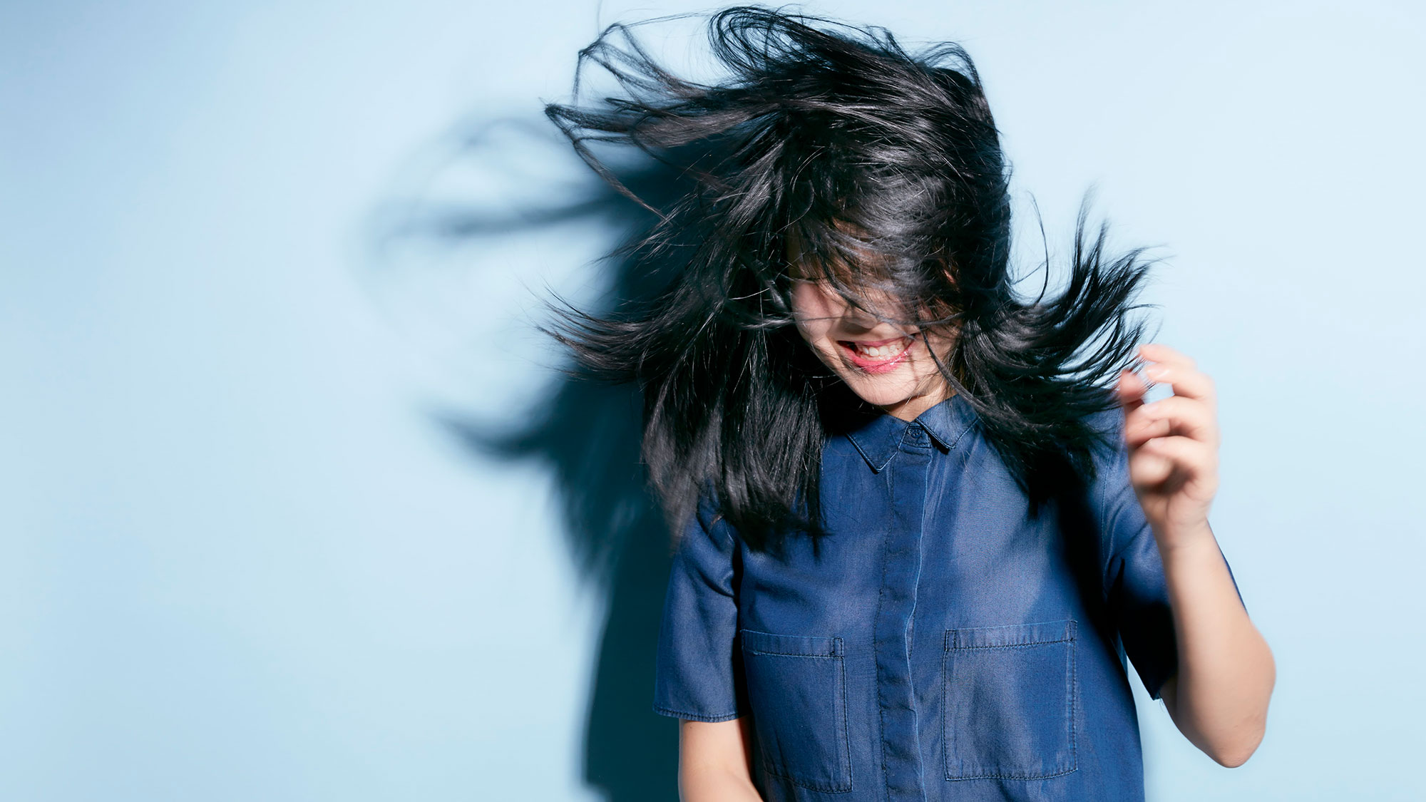 A young Asian Woman in a blue shirt shakes her long black hair, concealing much of her face