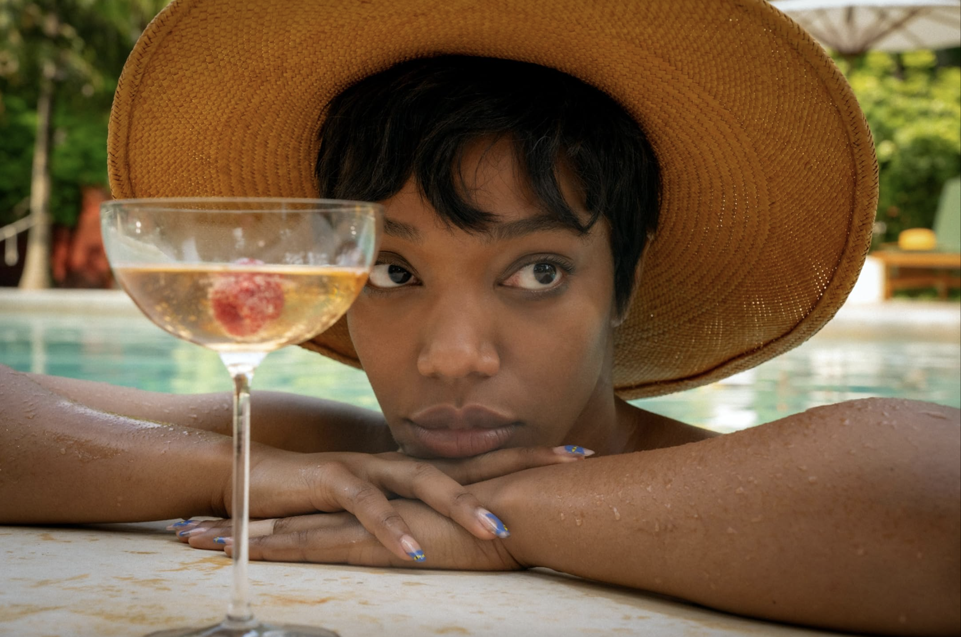 A pensive looking woman props herself up on the side of a pool directly behind a glass of champagne. She is wearing a sun hat.
