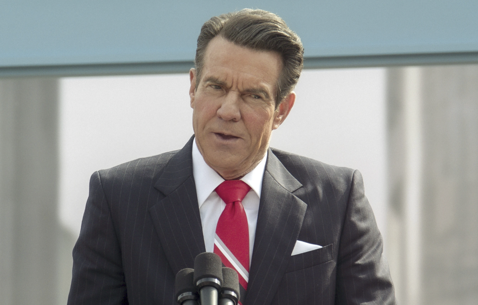 A white man with neat hair wearing a grey suit and red tie stands at a podium and speaks above three microphones.