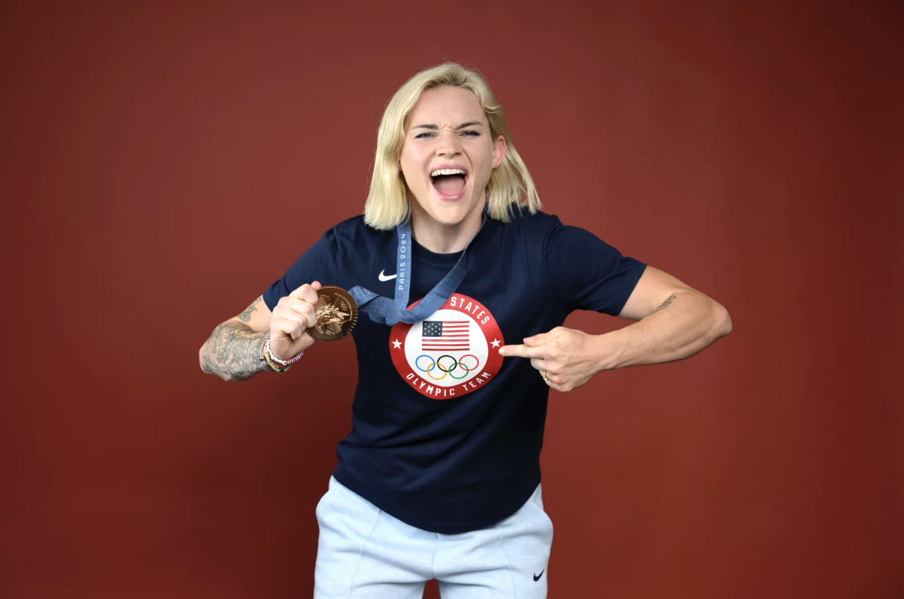 A blonde woman wearing an Olympic USA t-shirt points at a medal hanging around her neck and looks overjoyed.