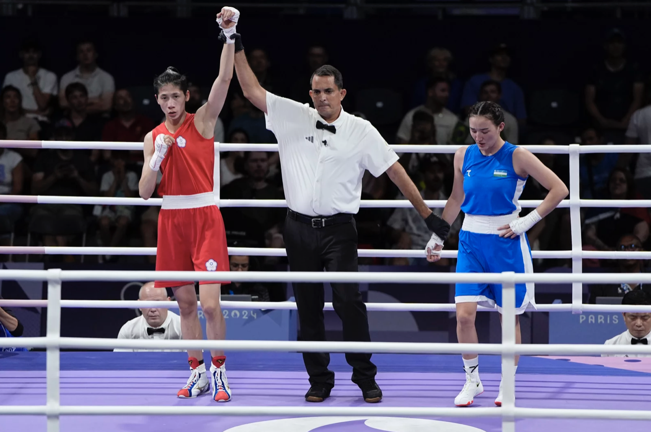 Inside a boxing ring, a referee holds aloft one arm of a female boxer dressed in red. A female boxer in blue looks down.