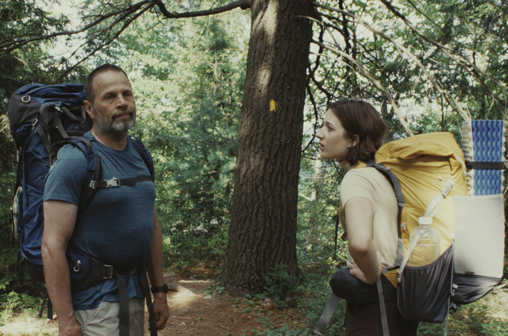 A middle-aged man and teenage girl stand facing each other in the woods, looking tired. Both are wearing large backpacks. 