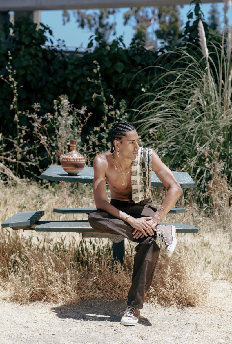 A shirtless man sits on a park bench with his right leg folded in his lap, looking off into the distance. 