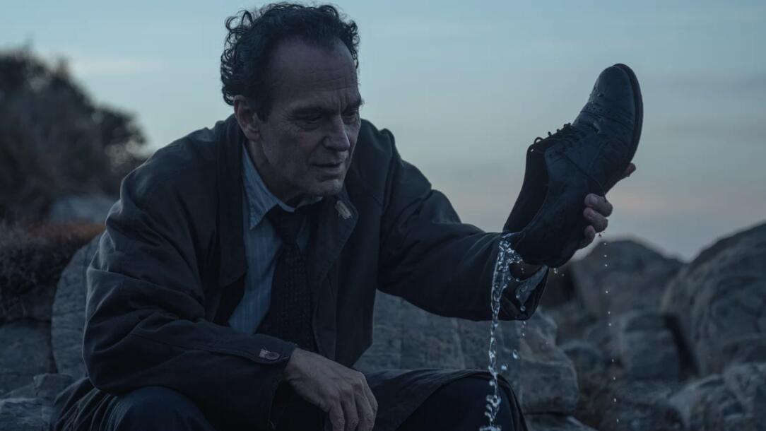 A man sits dejected outdoors, pouring water out of a leather shoe.