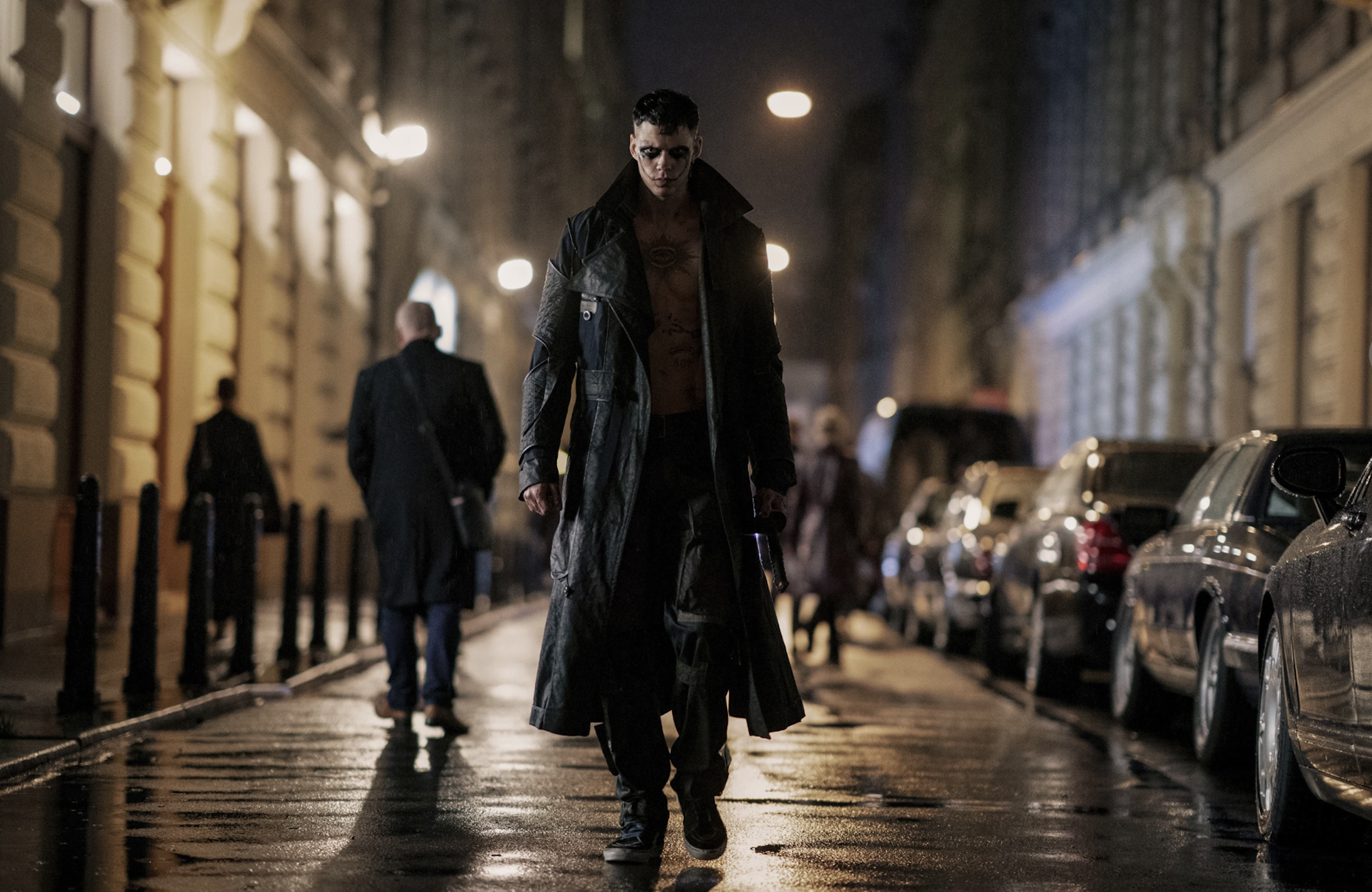 A man in a long black trench coat and gothic make up walks through damp city streets.