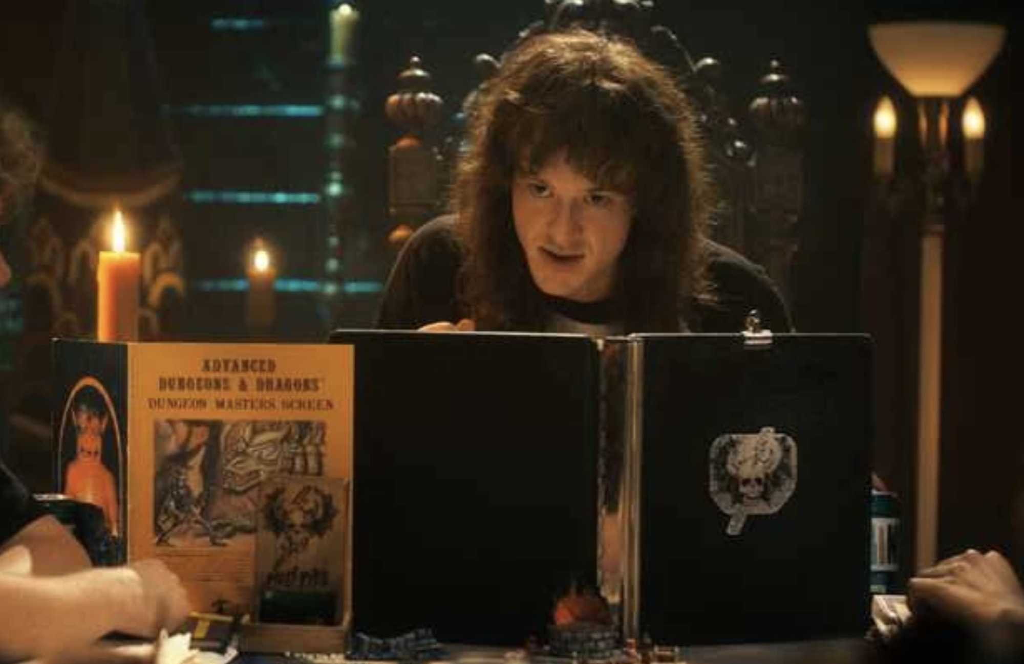 A man with long curly hair sits at a table behind large musty books.