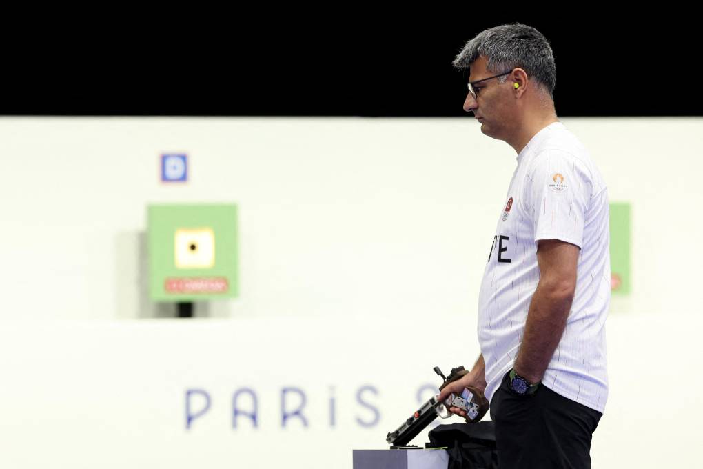 A man with one hand in his pocket, wearing a white t-shirt stands in front of a 'Paris' sign holding a gun.
