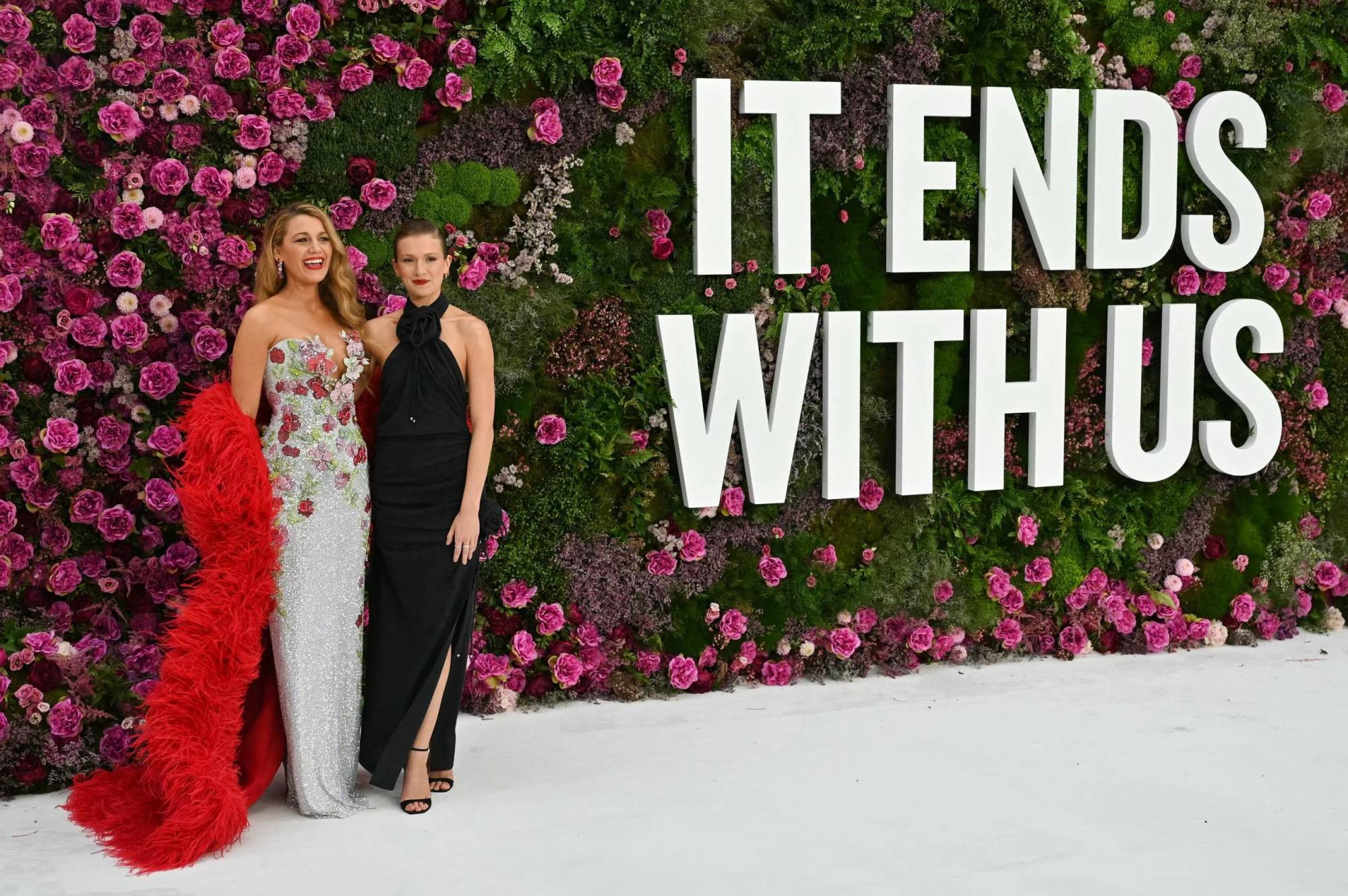 Two white women stand on a red carpet wearing gowns, next to lettering that reads ‘It Ends With Us.’