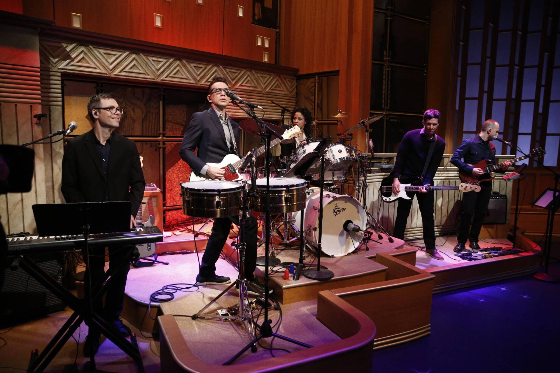 Four men and one woman wearing smart casual clothes play on a television studio stage.