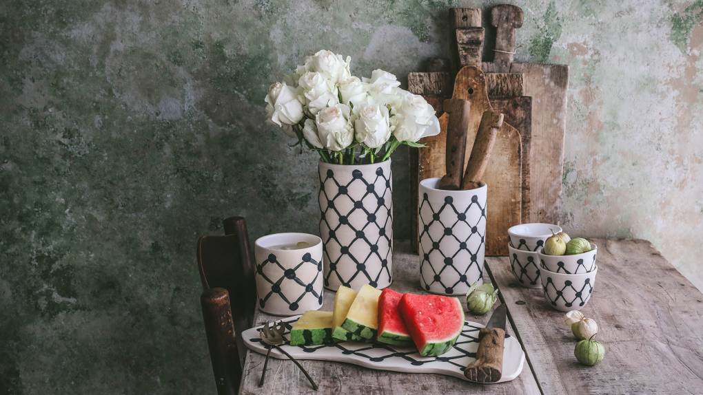 A tableau of ceramics with a keffiyeh-inspired checkerboard design, white roses and slices of watermelon, all on a rustic wooden table.