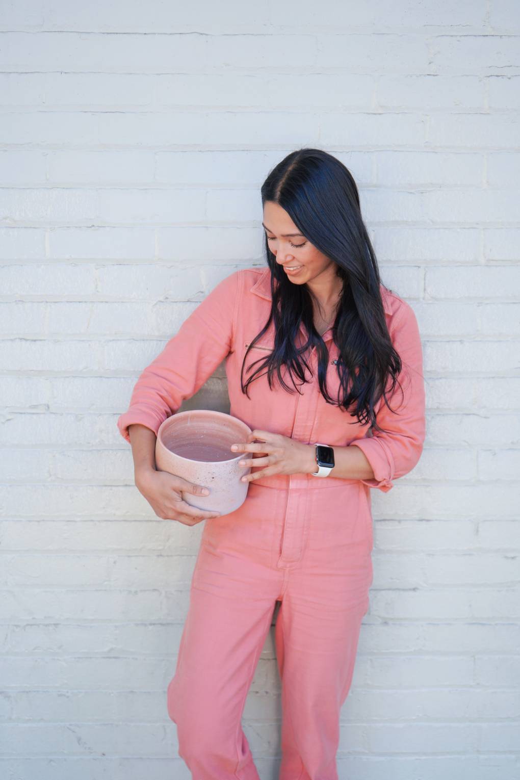 A ceramicist in a pink jumpsuit poses for a portrait while looking down at a large pink bowl.