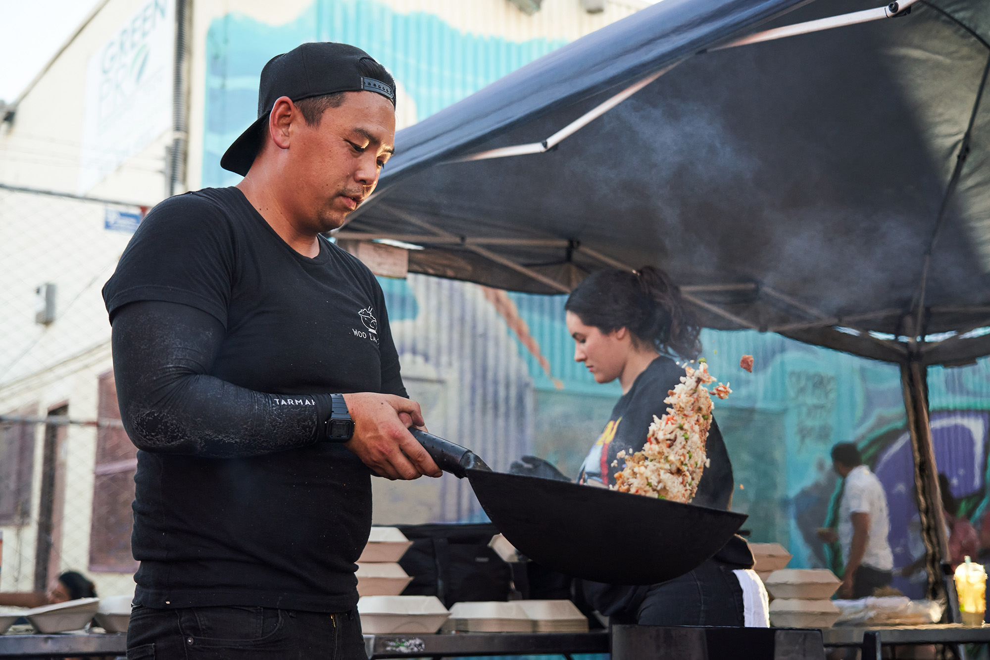 Asian man in black backwards baseball cap tosses fried rice in a hot wok at an outdoor market.