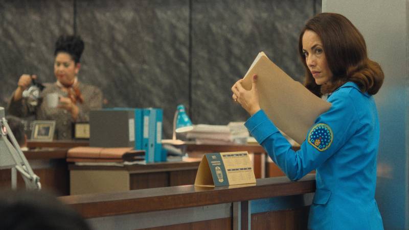 A woman in a 1960s-style blue dress sneaks a look inside a brown file filled with papers.