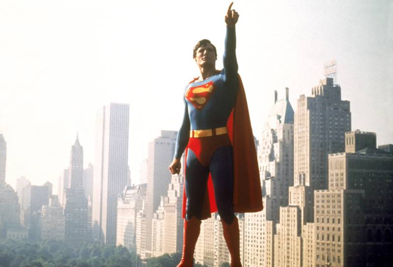 A man dressed as Superman points to the sky with a city skyline behind him.