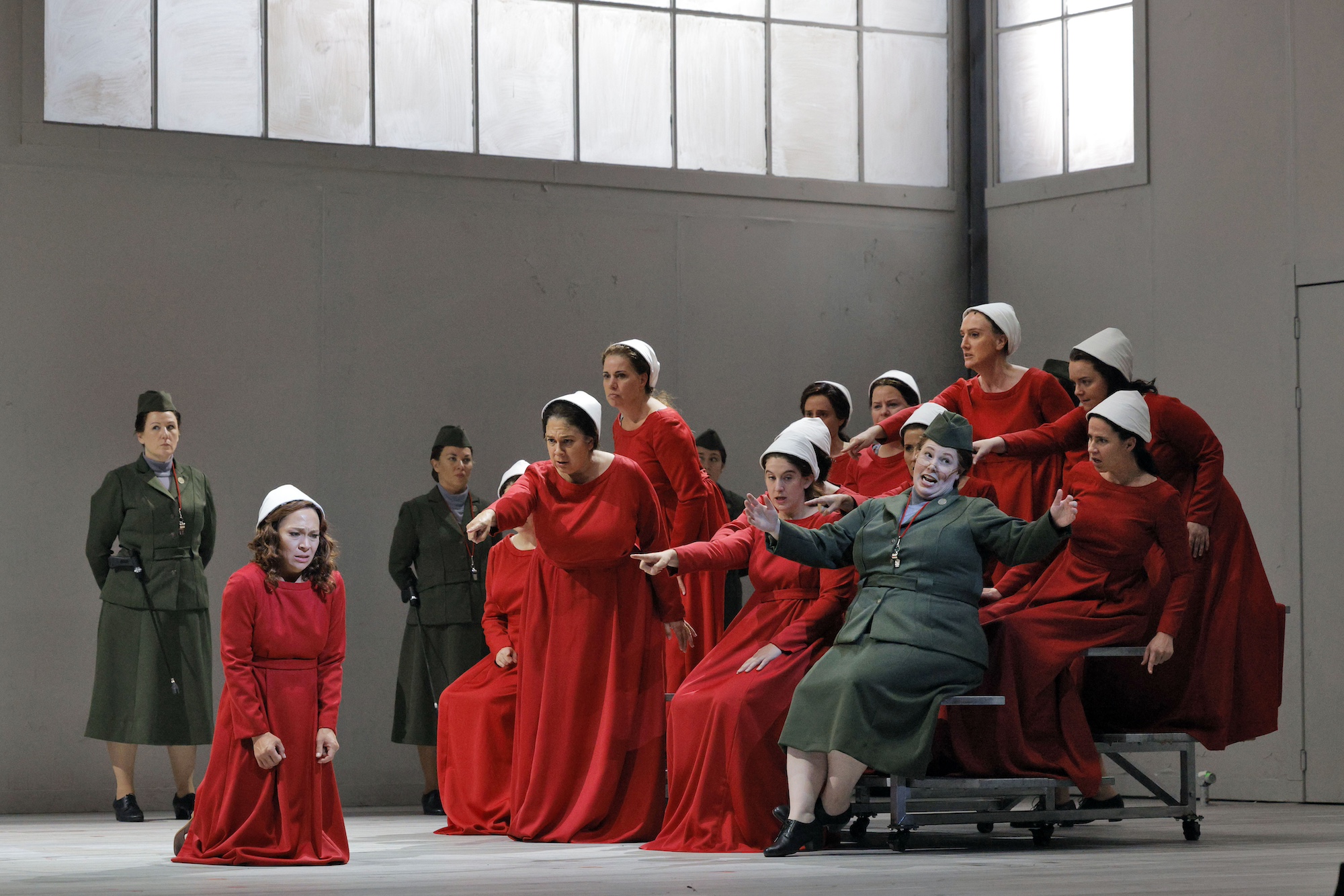 A woman kneels dejected on the floor as she is goaded and mocked by a group of other women. All but one are wearing red dresses and white bonnets.