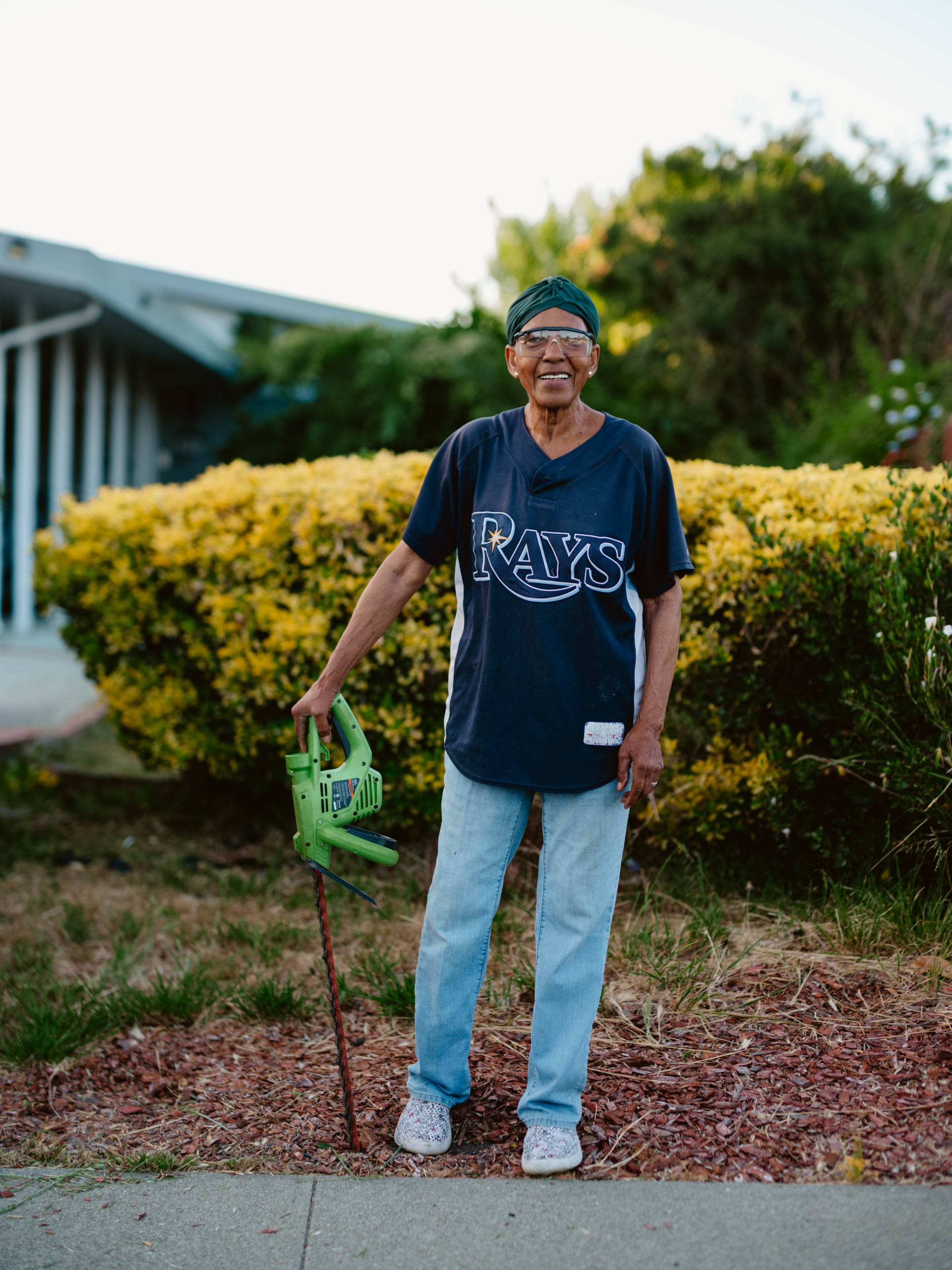 an elderly woman stands in front of her yard with a bush trimmer