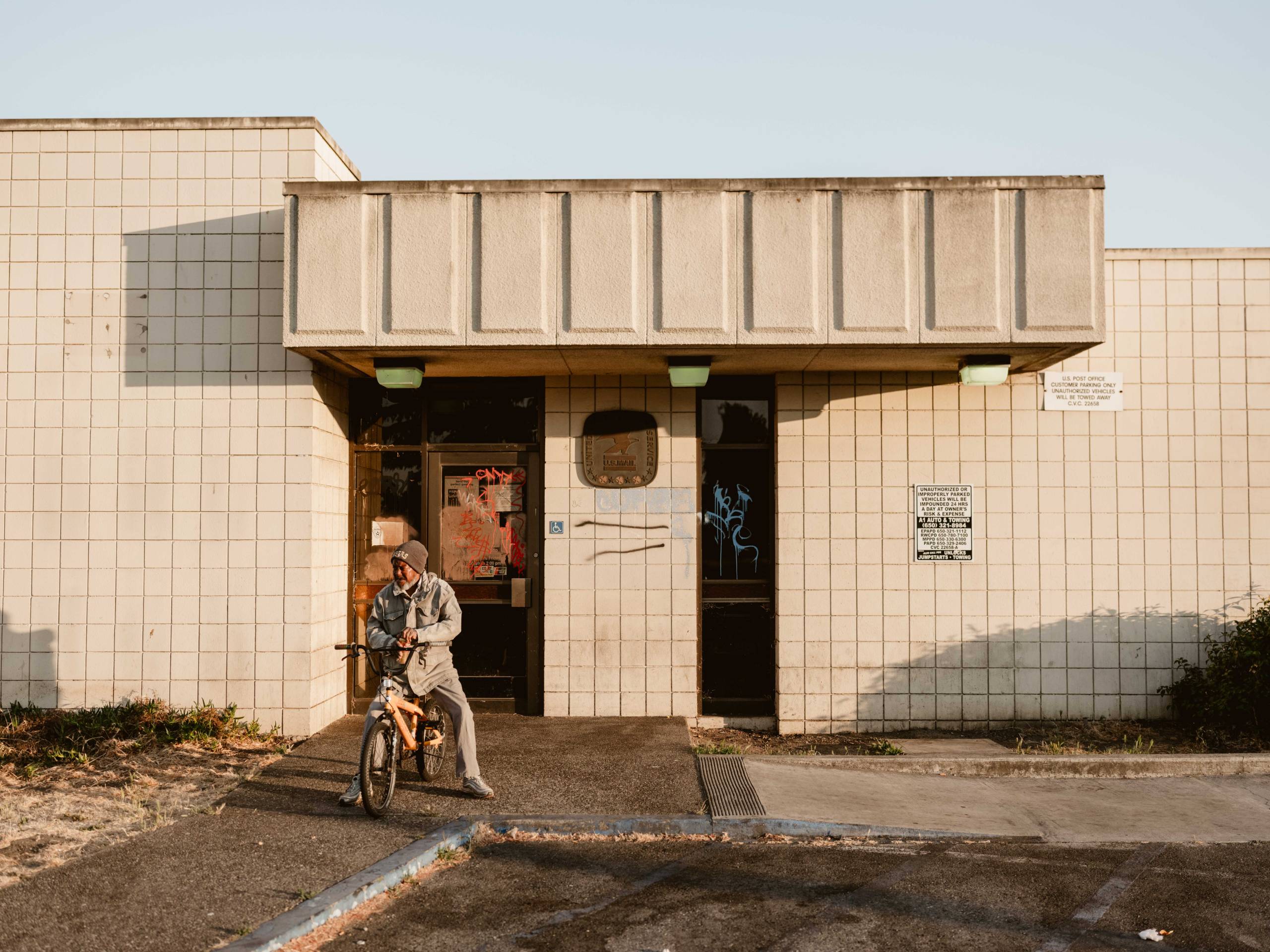 an unhoused man opens a can of beer in front of an abandoned post office in East Palo Alto