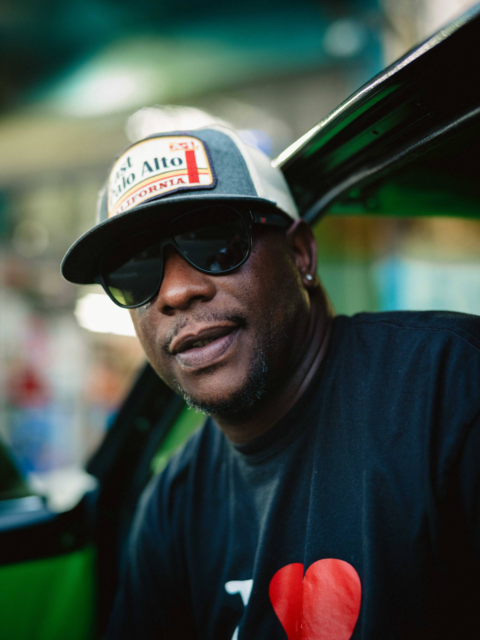 a profile shot of a man wearing an "I Love East Palo Alto" shirt and hat sits in his car with the door open in front of a liquor store